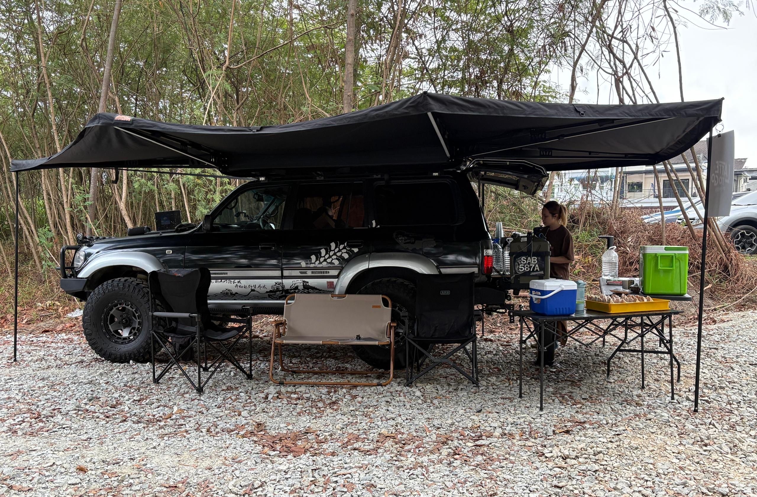 Black SUV with a large black awning set up for camping in a wooded area.